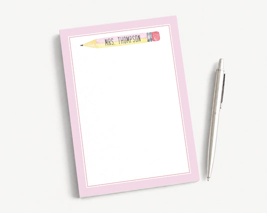 A pink-framed notepad with a pencil on top, accompanied by a pen, is placed on a white surface.