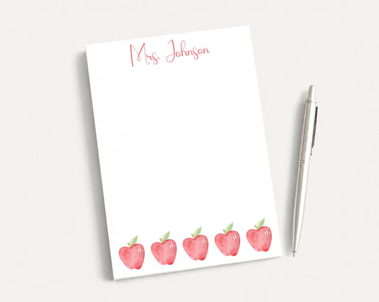 a white notepad with a watercolor illustration of five red apples, accompanied by a pen on a white surface.