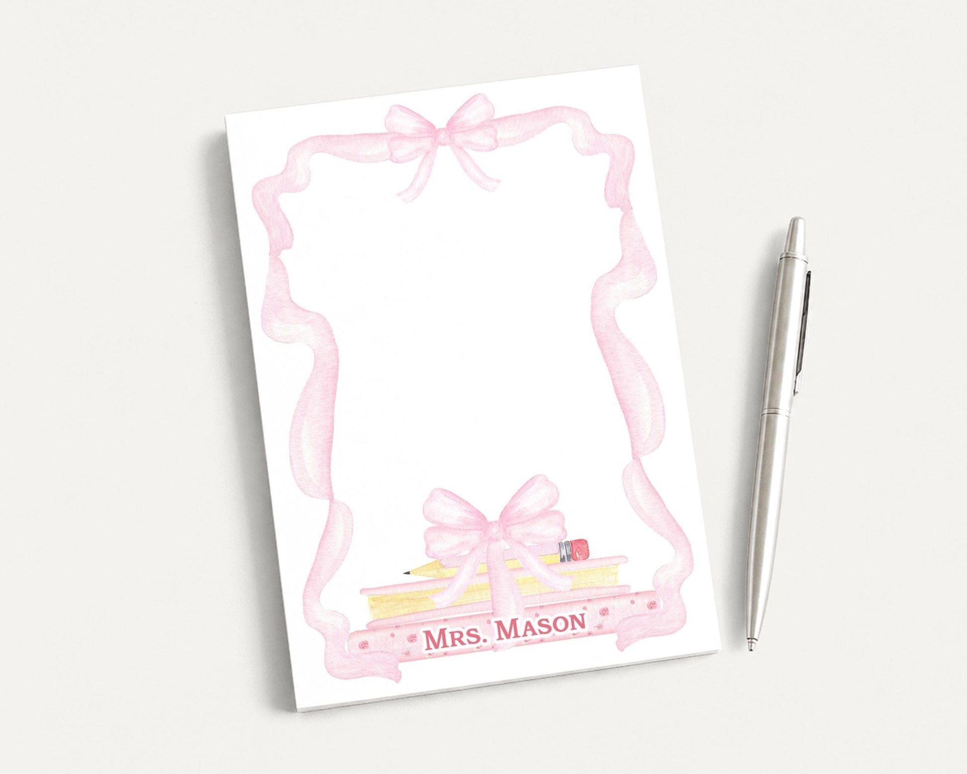 A white notepad with a pink ribbon frame and a pencil on top, along with a pen, is placed on a white surface.
