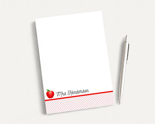 A white notepad with a red apple design and the name "Mrs. Henderson" printed on it, along with a silver pen, are placed on a white surface.