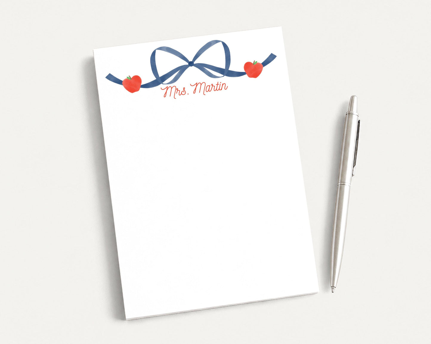 A blank white notepad with a blue ribbon and two red hearts on it, accompanied by a silver pen on a plain white background.