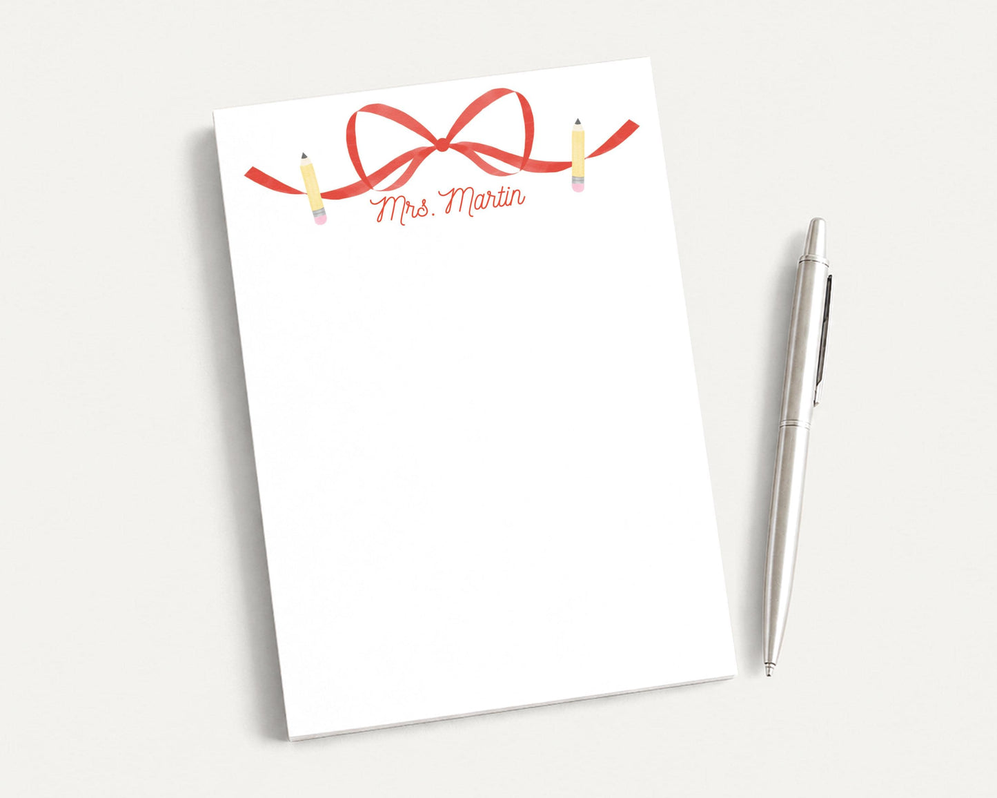 a blank white notepad with a red ribbon and pencils on it, placed next to a silver pen on a white surface.
