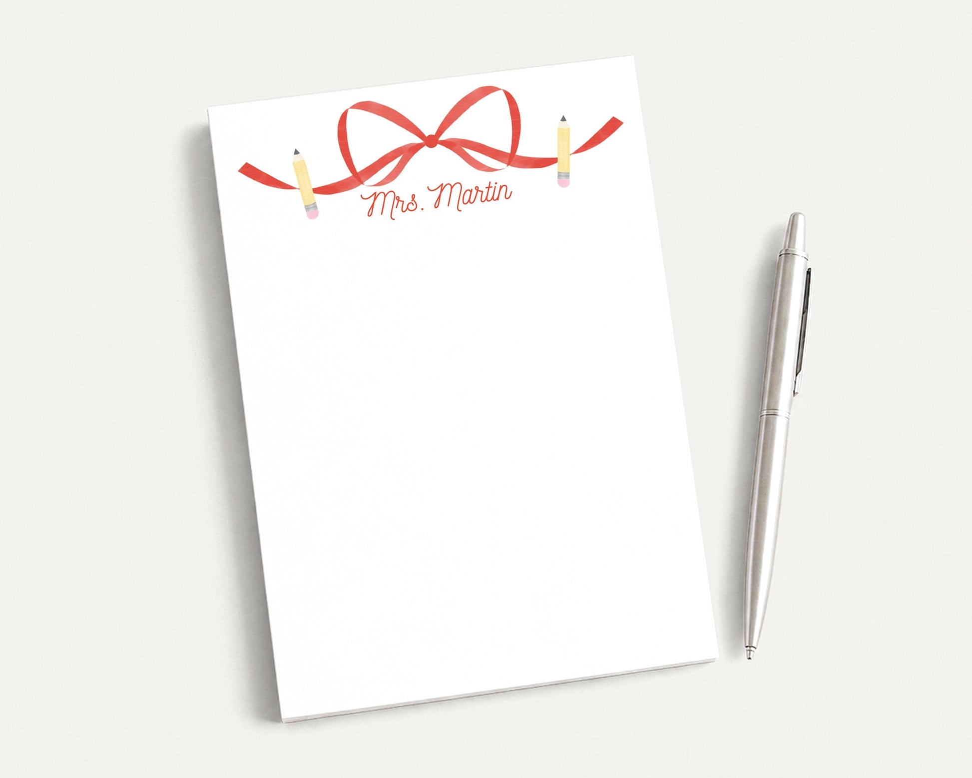 a blank white notepad with a red ribbon and pencils on it, placed next to a silver pen on a white surface.