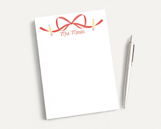 a blank white notepad with a red ribbon and pencils on it, placed next to a silver pen on a white surface.