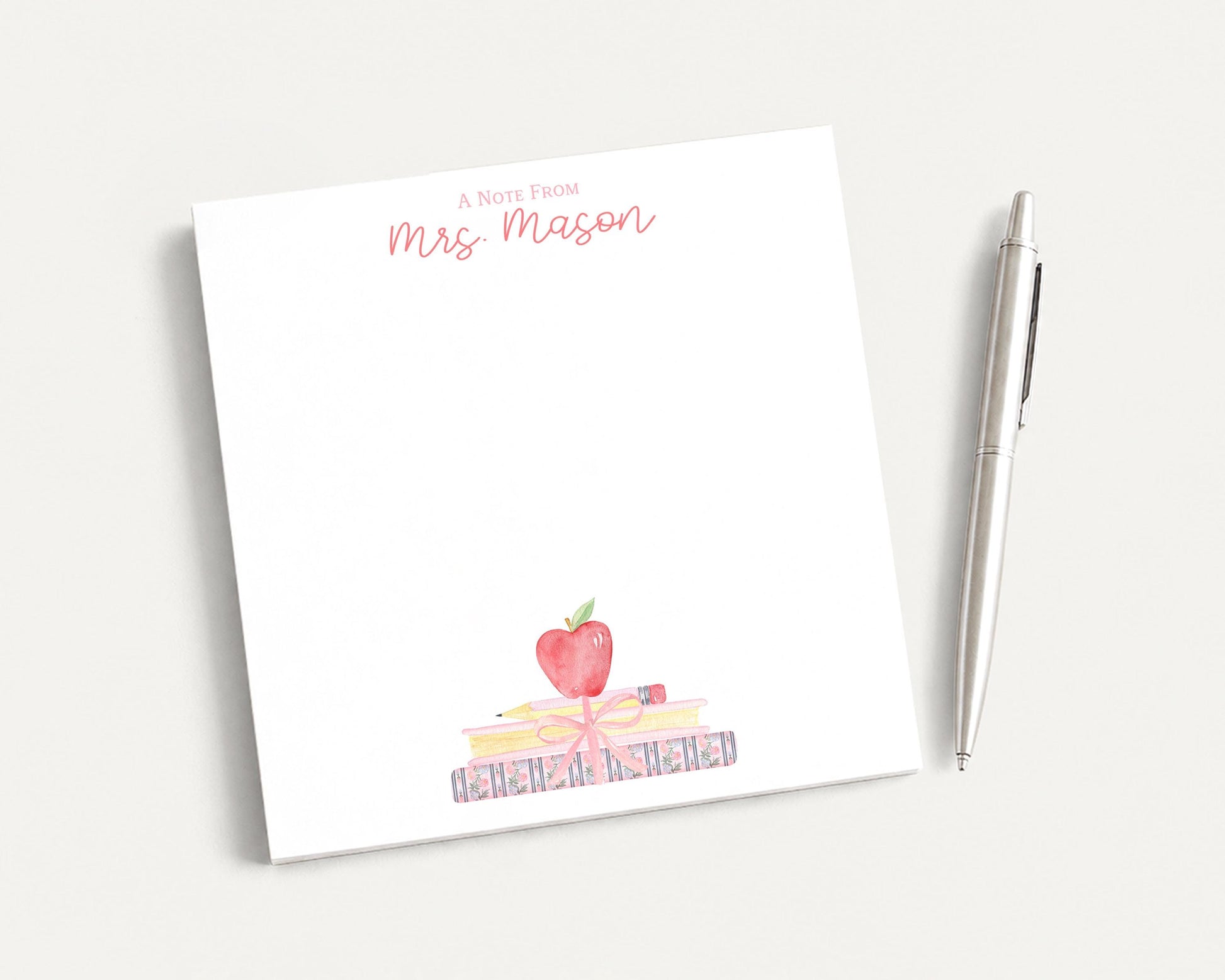 A white sticky note with a watercolor illustration of a stack of books and an apple on it, along with the text "A Note From Mrs. Mason" written on it. There is also a pen next to the sticky note.