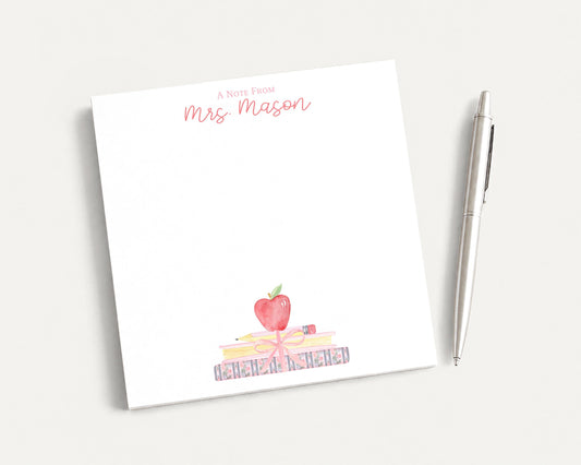 A white sticky note with a watercolor illustration of a stack of books and an apple on it, along with the text "A Note From Mrs. Mason" written on it. There is also a pen next to the sticky note.