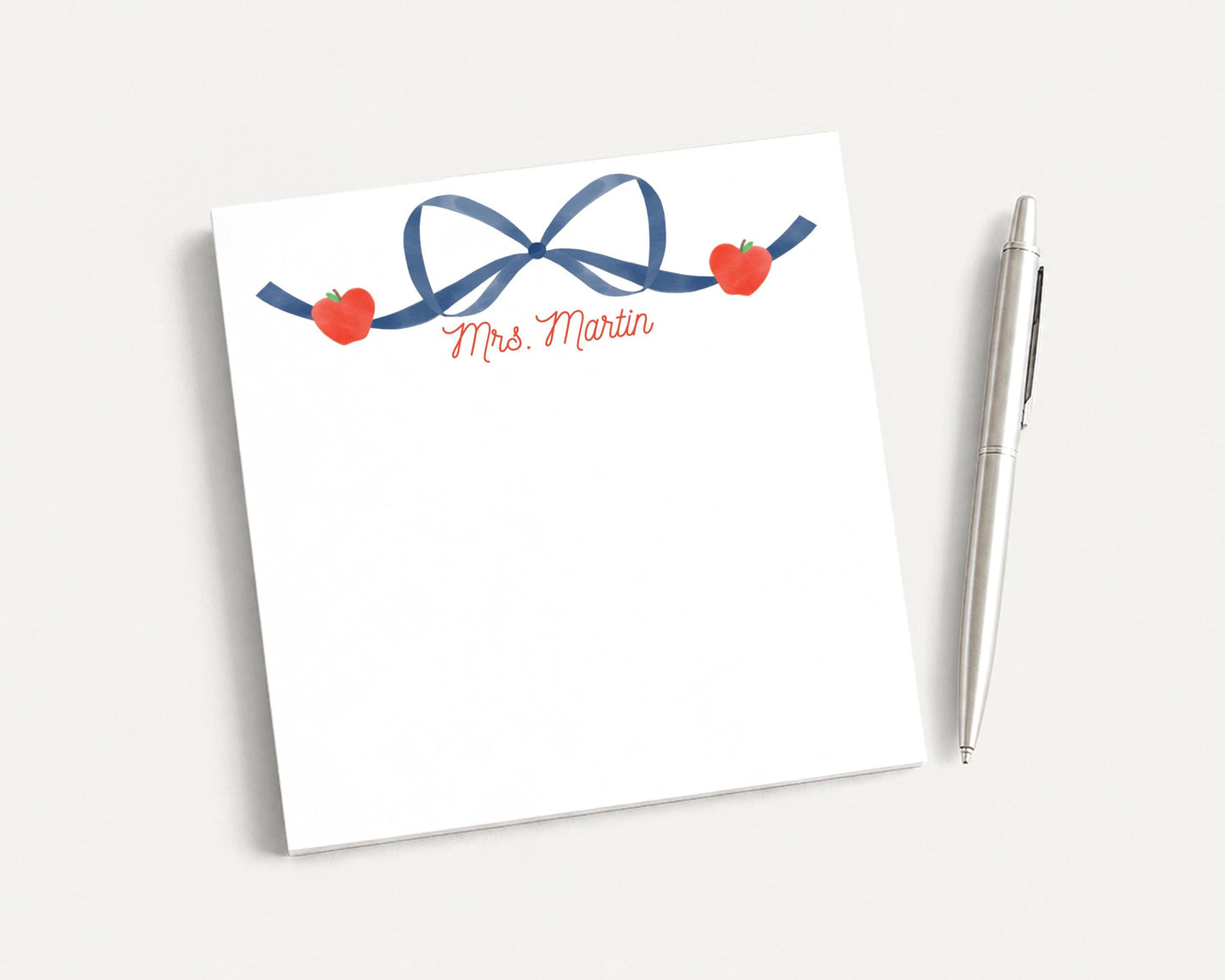 A white notepad with a blue ribbon and two red apples, along with a pen, is placed on a white surface.