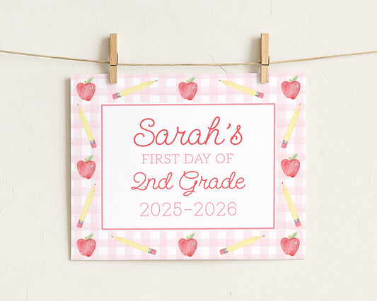 A pink and white sign with the text "Sarah's FIRST DAY OF 2nd Grade" and "2025-2026" printed on it, along with illustrations of pencils and apples.