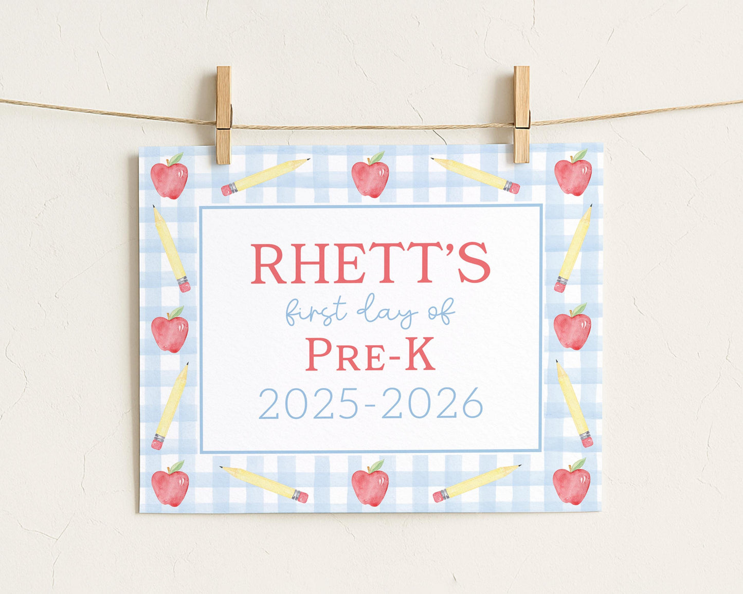 A sign with the text "RHETT'S first day of PRE-K" and "2025-2026" printed on it, surrounded by illustrations of pencils and apples.