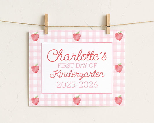 A pink and white checkered sign with the text "Charlotte's first day of Kindergarten 2025-2026" and several red apples on a string.