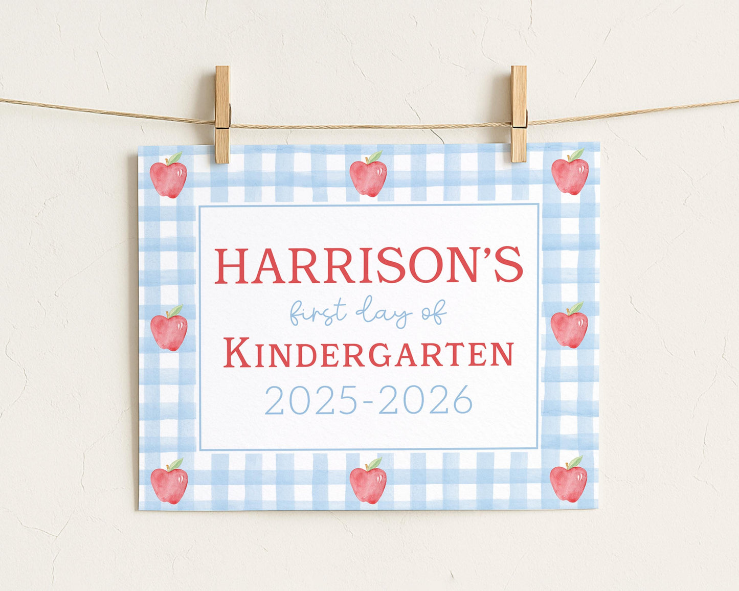 A sign with the text "HARRISON'S first day of Kindergarten" and "2025-2026" printed on it, along with a blue and white checkered pattern and red apples.
