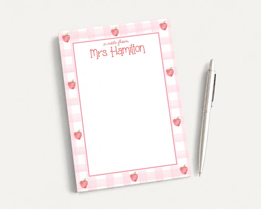 A pink and white checkered notepad with a blank page and a pen on a white surface.