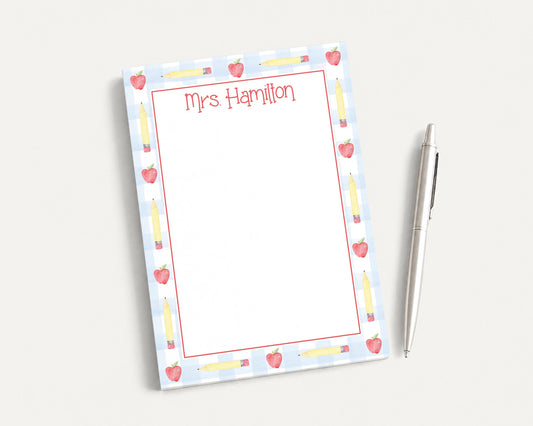 A personalized nameplate with a pencil and apple design, placed next to a pen on a white surface.