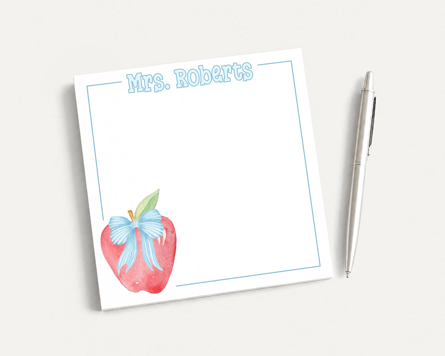 a white sticky note with a blue border, featuring an illustration of an apple with a bow on it. Next to the sticky note is a pen, suggesting that it is being used to write on the note.
