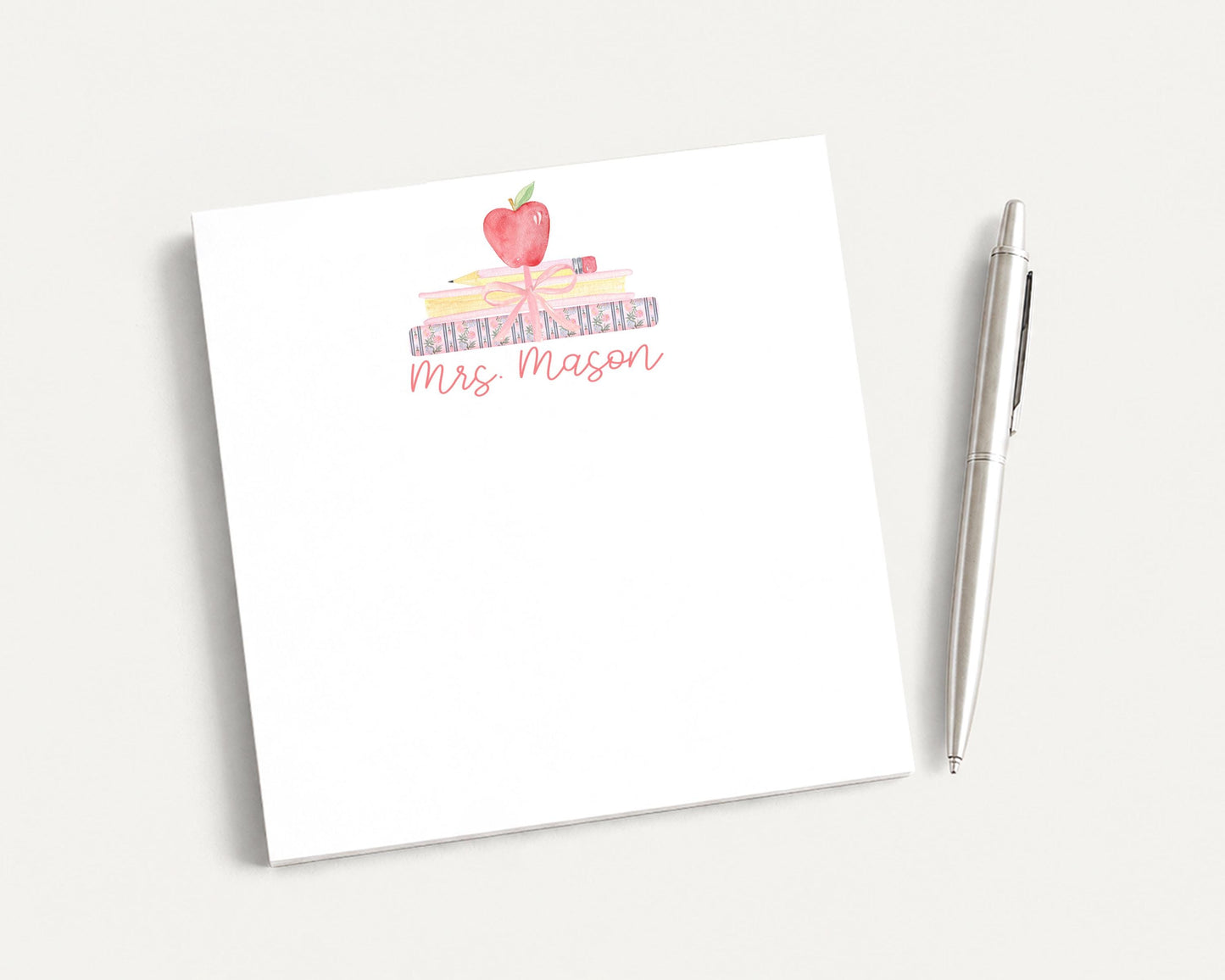 A white notepad with a watercolor illustration of an apple and the text "Mrs. Mason" on it, along with a pen on a plain white surface.