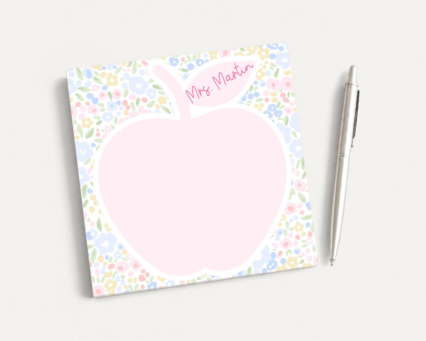 A pink flower-patterned notepad with a blank space in the center, accompanied by a silver pen on a plain white background.