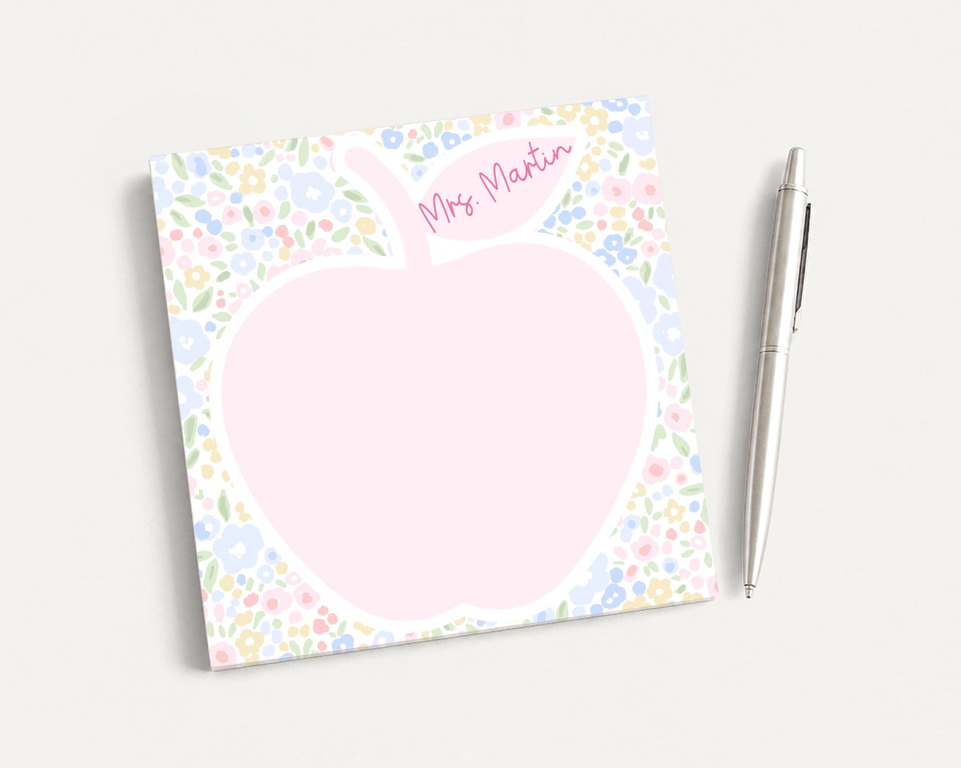 A pink flower-patterned notepad with a blank space in the center, accompanied by a silver pen on a plain white background.