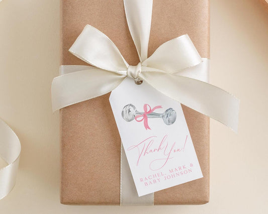 A brown gift box with a white ribbon and a white tag attached to it. The tag has a pink ribbon and a drawing of a dumbbell, and it says "Thank you! Rachel Mark & Baby Johnson".