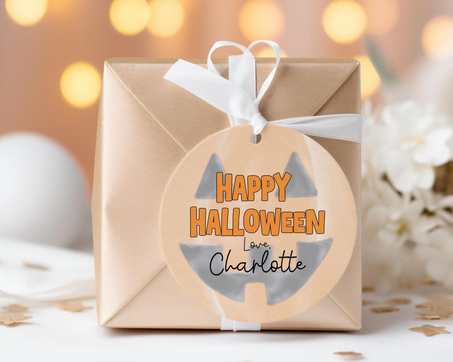 A brown gift box with a Halloween-themed tag that reads "Happy Halloween Love Charlotte" is displayed on a table with white flowers in the background.