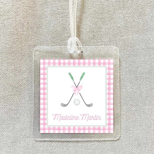 A clear plastic name tag with a pink and white checkered border, featuring a golf club and ball graphic, and the name "Madeeline Martin" written below.