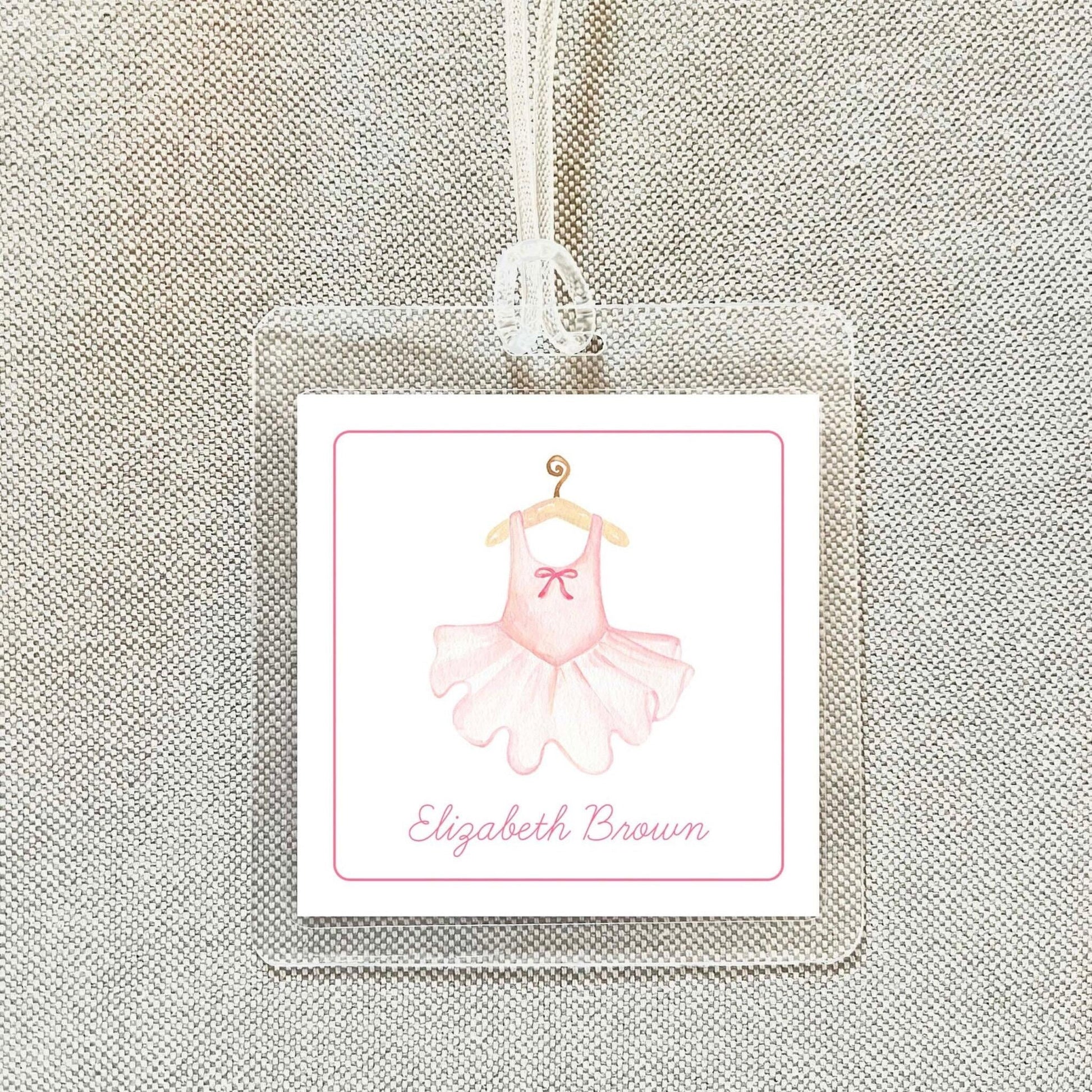 A clear plastic tag with a pink tutu and bow on it, hanging from a string.