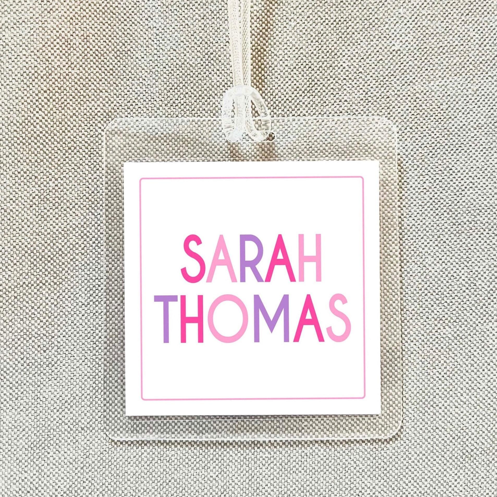 A clear plastic name tag with the name "SARAH THOMAS" printed on it in pink and purple colors.