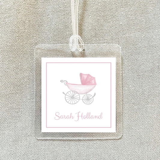 A clear plastic tag with a pink stroller and the name "Sarah Holland" printed on it, hanging on a fabric background.