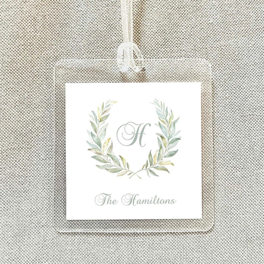 A clear plastic name tag with a monogrammed "H" and a wreath design, hanging on a beige fabric background.