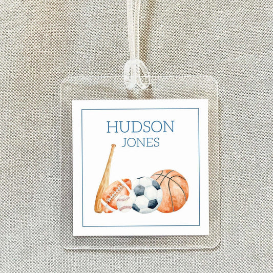 A clear plastic name tag with a sports equipment design, including a baseball bat, a football, and a basketball, attached to a string.