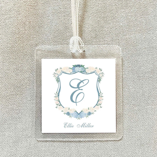A clear plastic tag with a monogrammed shield design and floral elements, hanging on a beige fabric background.