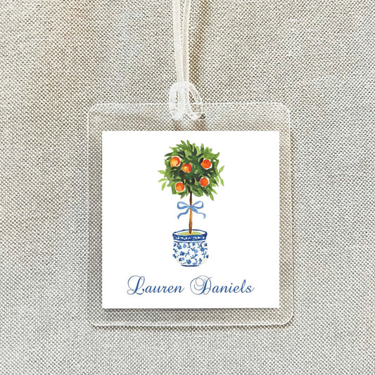 A square tag with a watercolor illustration of a potted tree with oranges on top, hanging from a string.