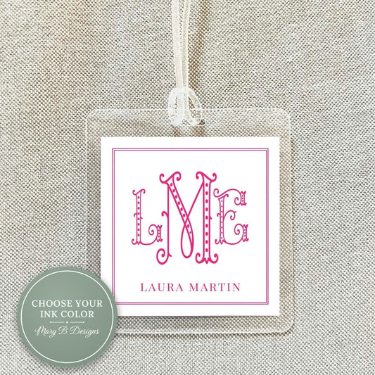 A clear plastic tag with a pink monogram and text, hanging on a fabric background.