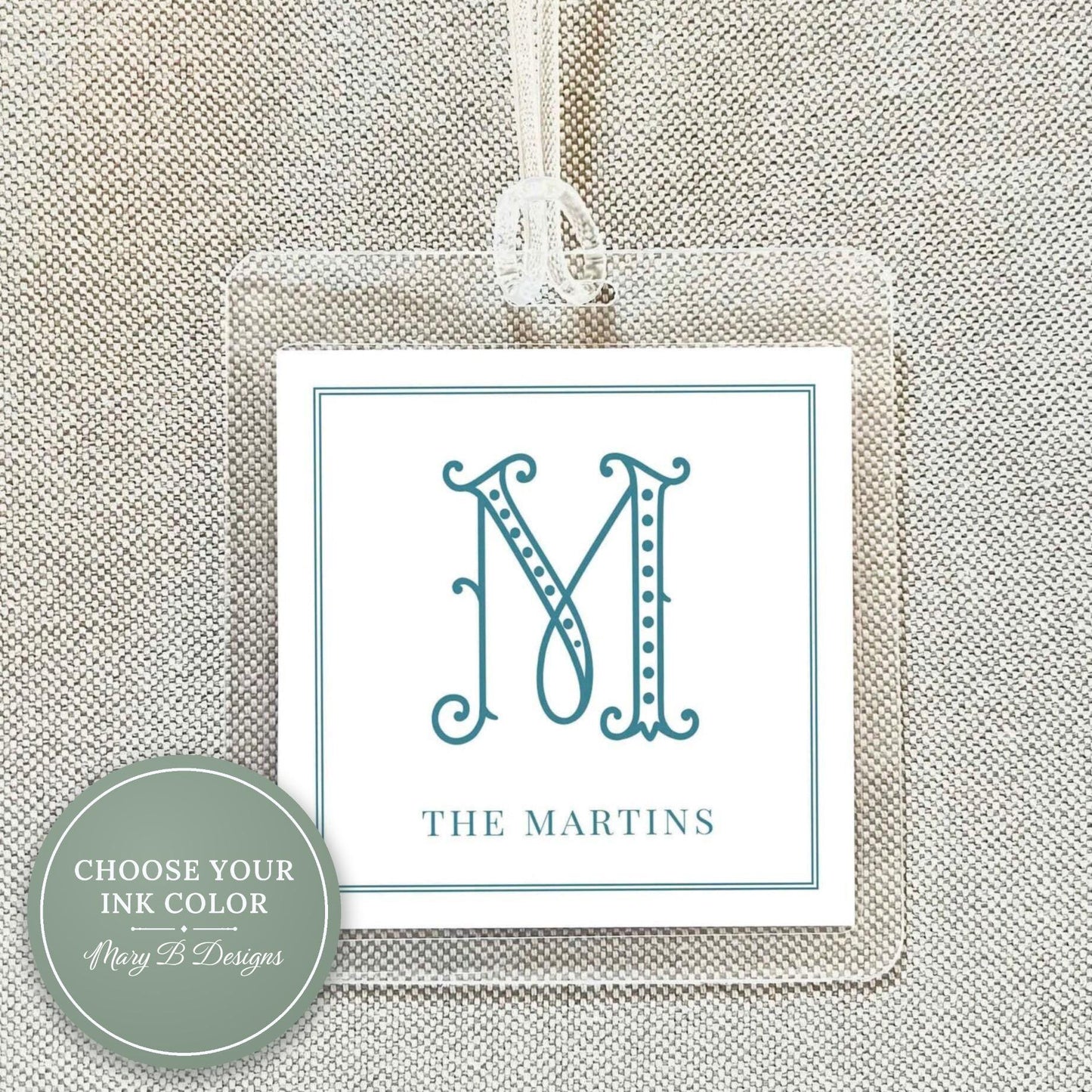 May include: Clear acrylic luggage tag with a white insert featuring a teal letter "M" in an elegant font. The text "THE MARTINS" is below the letter. A white ribbon is attached for hanging.