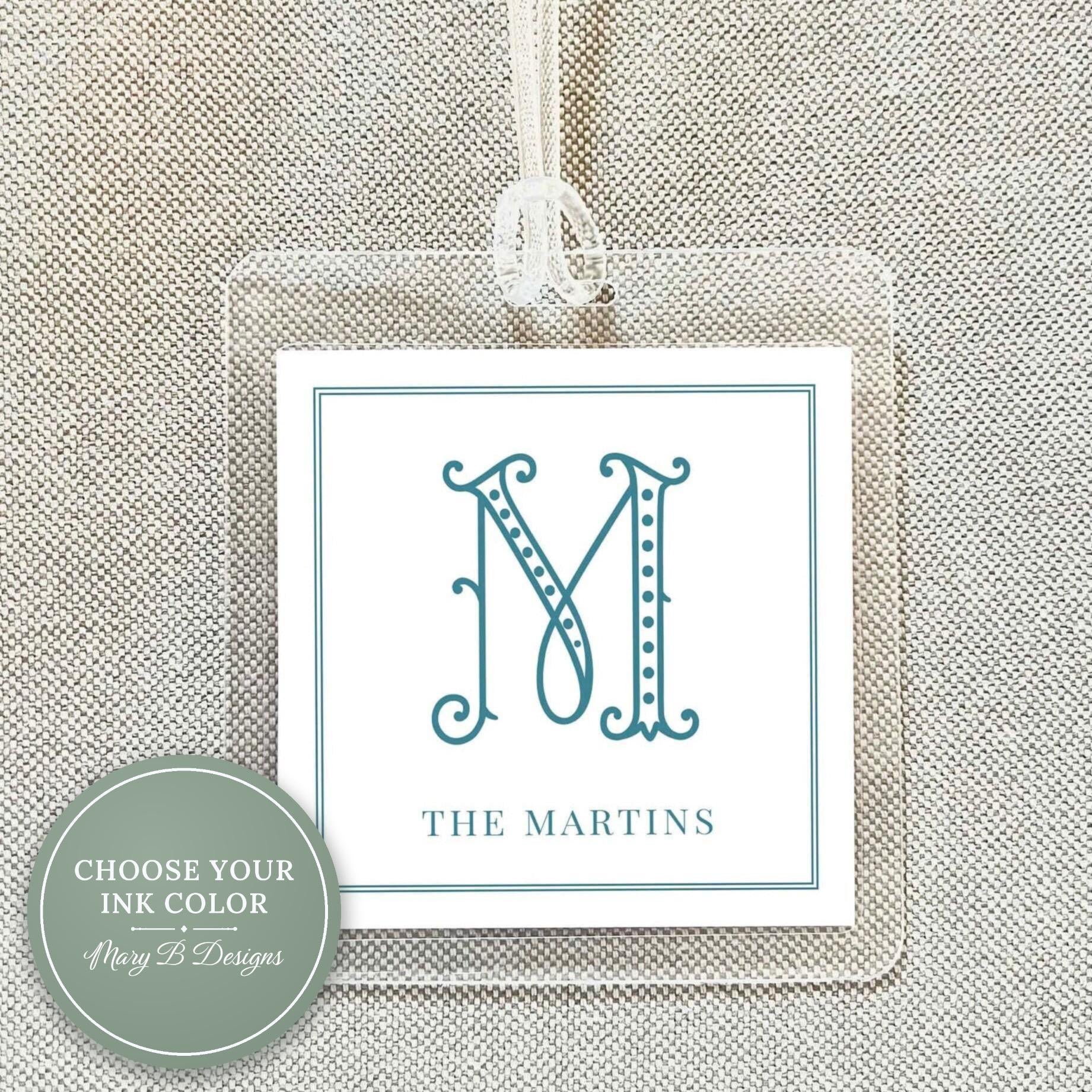 May include: Clear acrylic luggage tag with a white insert featuring a teal letter "M" in an elegant font. The text "THE MARTINS" is below the letter. A white ribbon is attached for hanging.