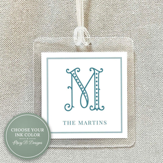 May include: Clear acrylic luggage tag with a white insert featuring a teal letter "M" in an elegant font. The text "THE MARTINS" is below the letter. A white ribbon is attached for hanging.