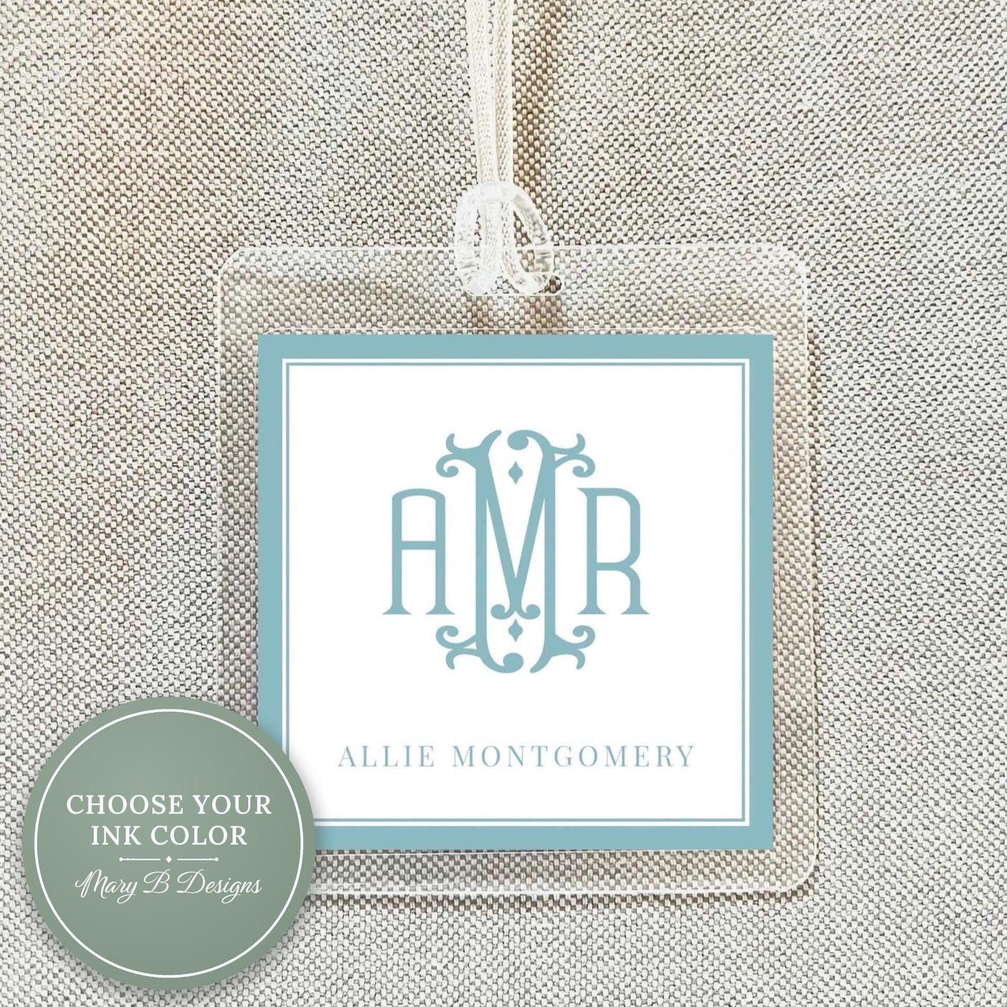 a clear luggage tag with a monogrammed initials logo, hanging on a beige fabric background.