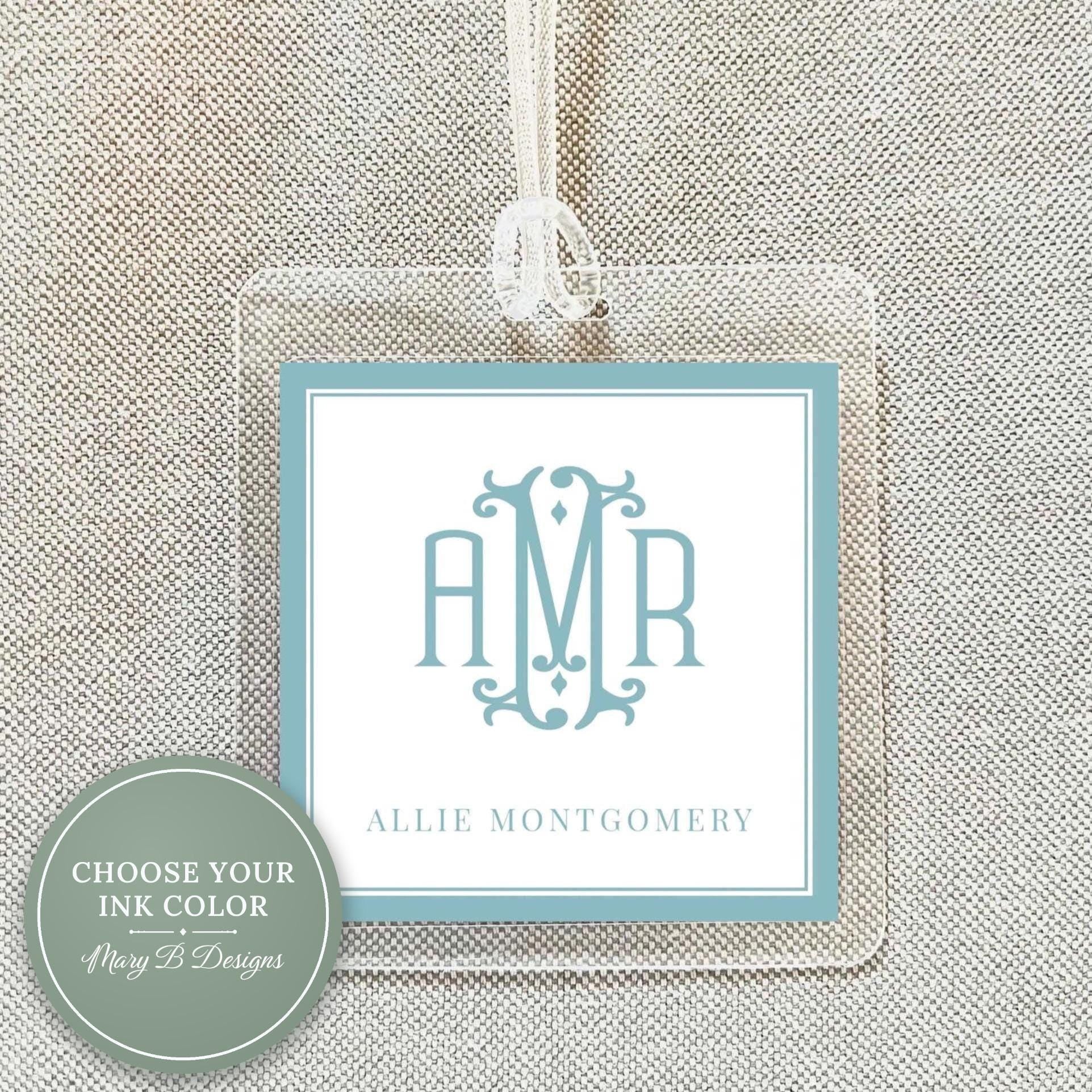 a clear luggage tag with a monogrammed initials logo, hanging on a beige fabric background.