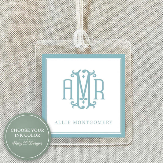 a clear luggage tag with a monogrammed initials logo, hanging on a beige fabric background.