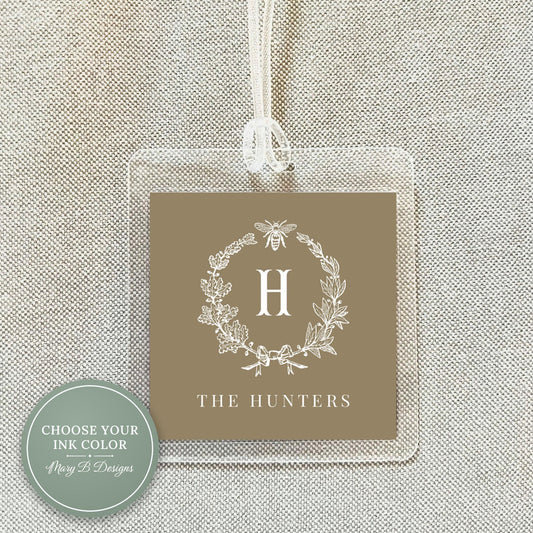 A clear plastic tag with a brown background and a white floral wreath design, featuring the letter "H" in the center.