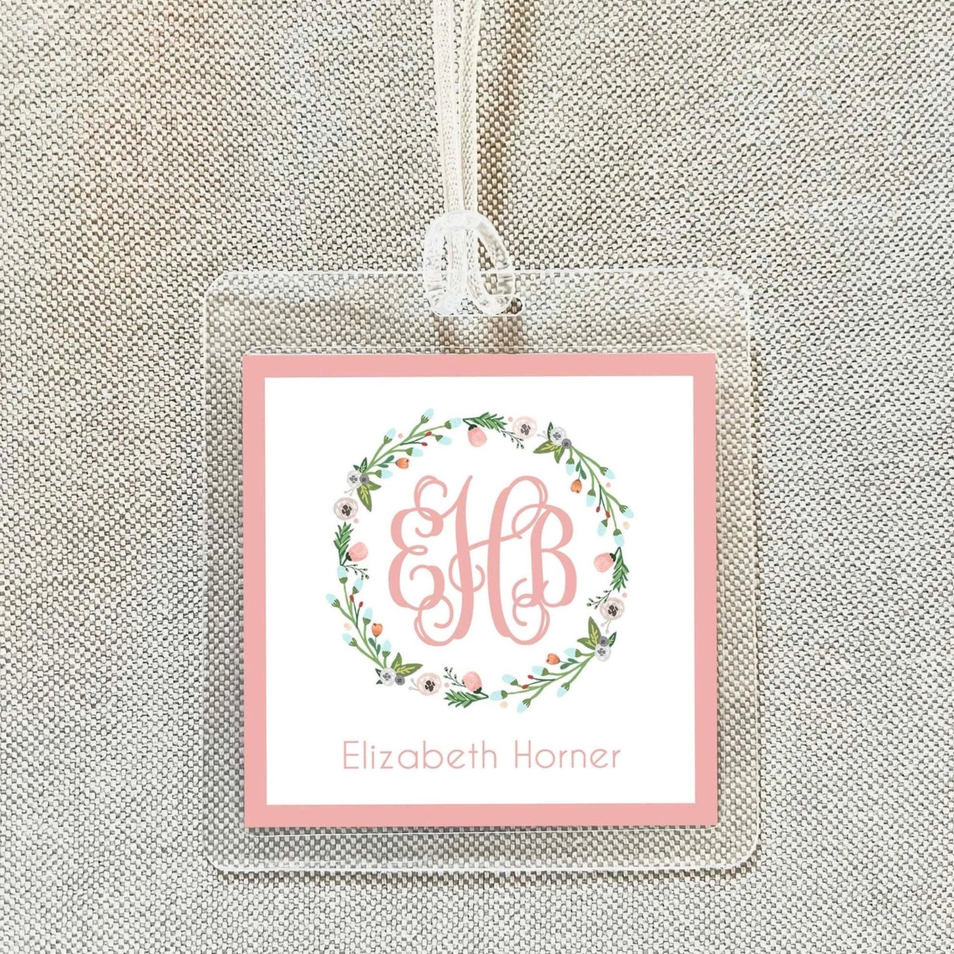A clear plastic tag with a pink border and a floral wreath design, featuring the initials "EB" in the center.