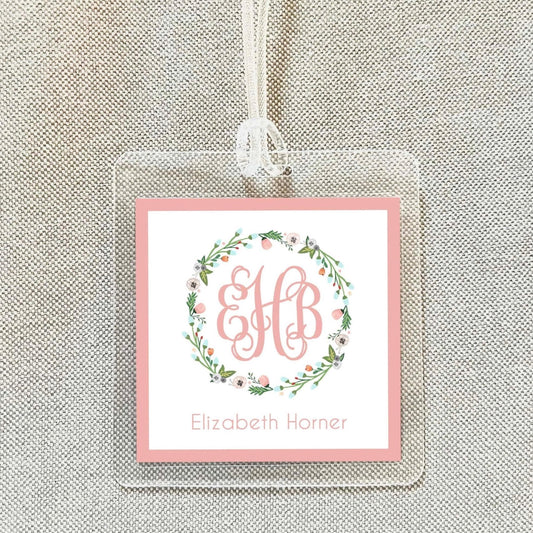 A clear plastic tag with a pink border and a floral wreath design, featuring the initials "EB" in the center.