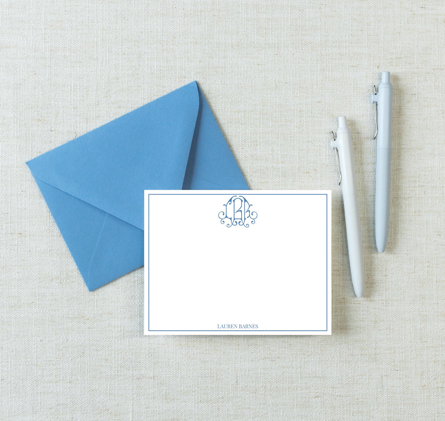 a blue envelope, a blank white card with a monogram, and two pens on a light-colored surface.