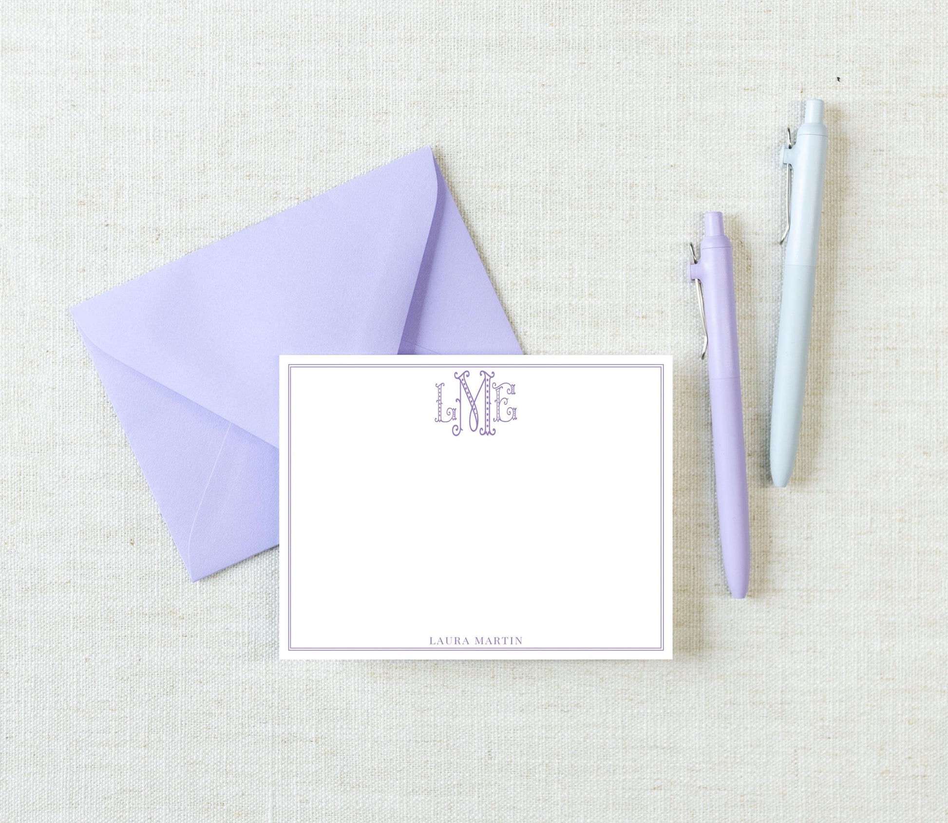 a purple envelope, a blank white card with a monogram, and two pens on a light-colored surface.