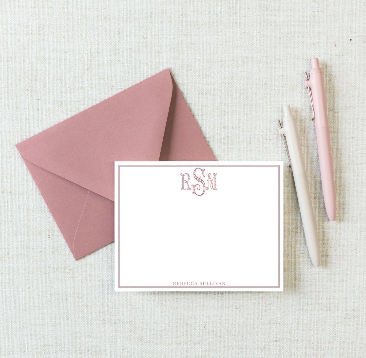 a pink envelope, a blank white card with a monogrammed logo, and two pens on a light-colored surface.