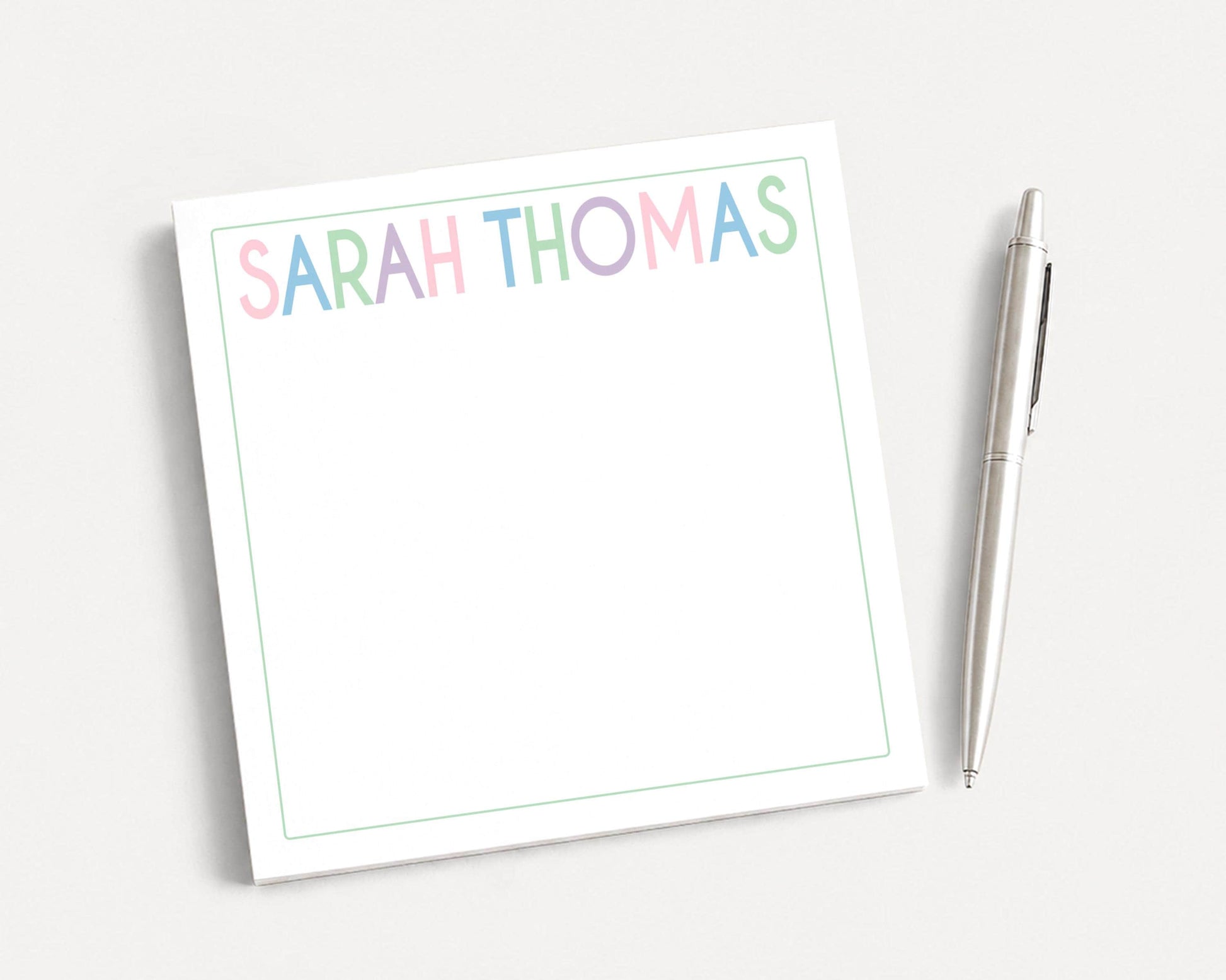 A white notepad with the name "SARAH THOMAS" written on it, along with a pen, is placed on a plain white surface.