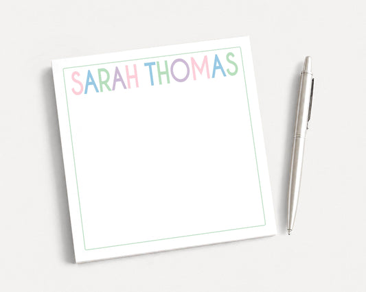 A white notepad with the name "SARAH THOMAS" written on it, along with a pen, is placed on a plain white surface.