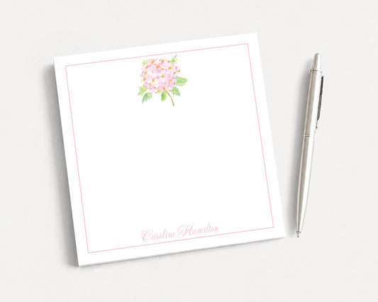 a white notepad with a pink floral design and a pen placed next to it on a white surface.