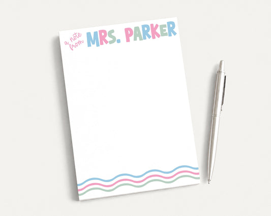 A blank notepad with the name "Mrs. Parker" written on it, along with a pen, is placed on a white surface.
