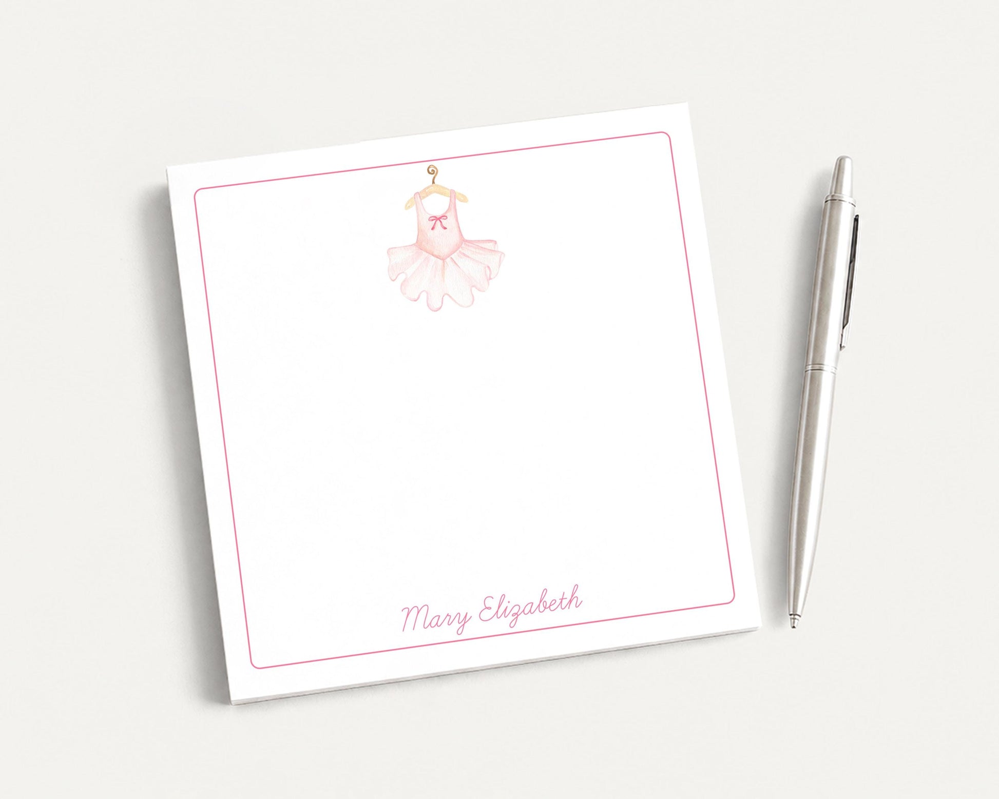 A white notepad with a pink ballet tutu illustration and the name "Mary Elizabeth" written on it, along with a pen on a plain white background.
