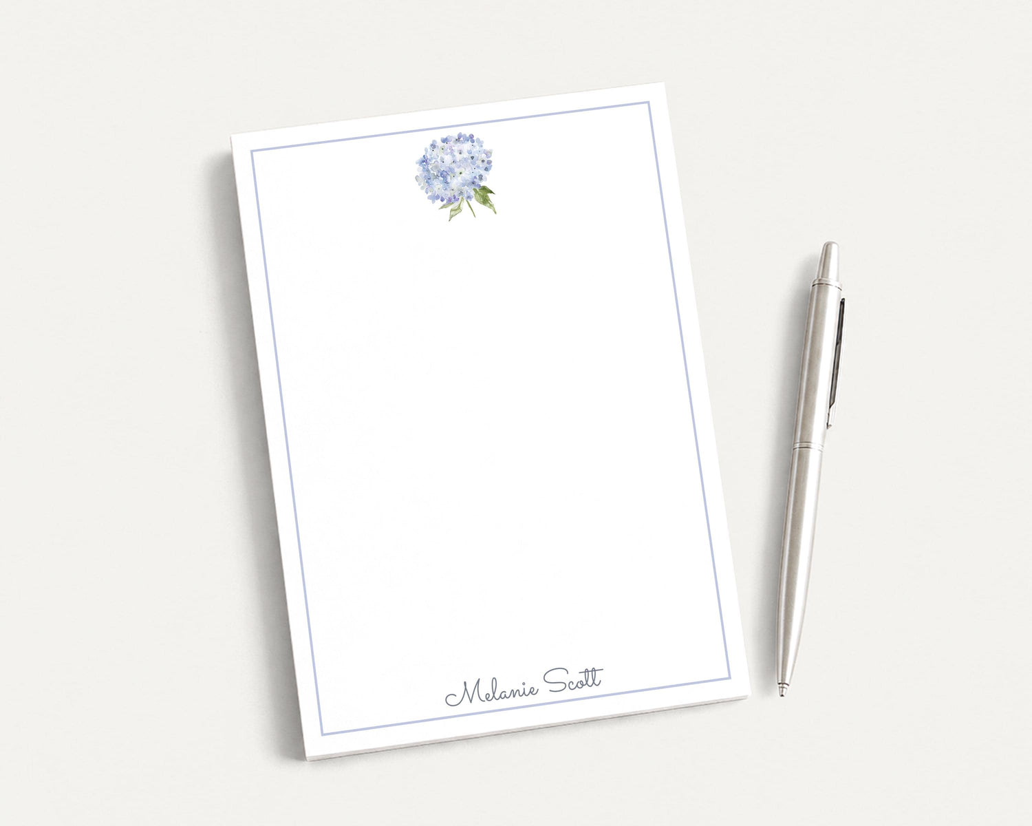 a blank white notepad with a blue border, and a silver pen is placed next to it on a white surface.